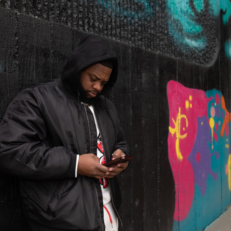 Young man wearing a hoodie looks at a cell phone leaning against a wall with graffitti.